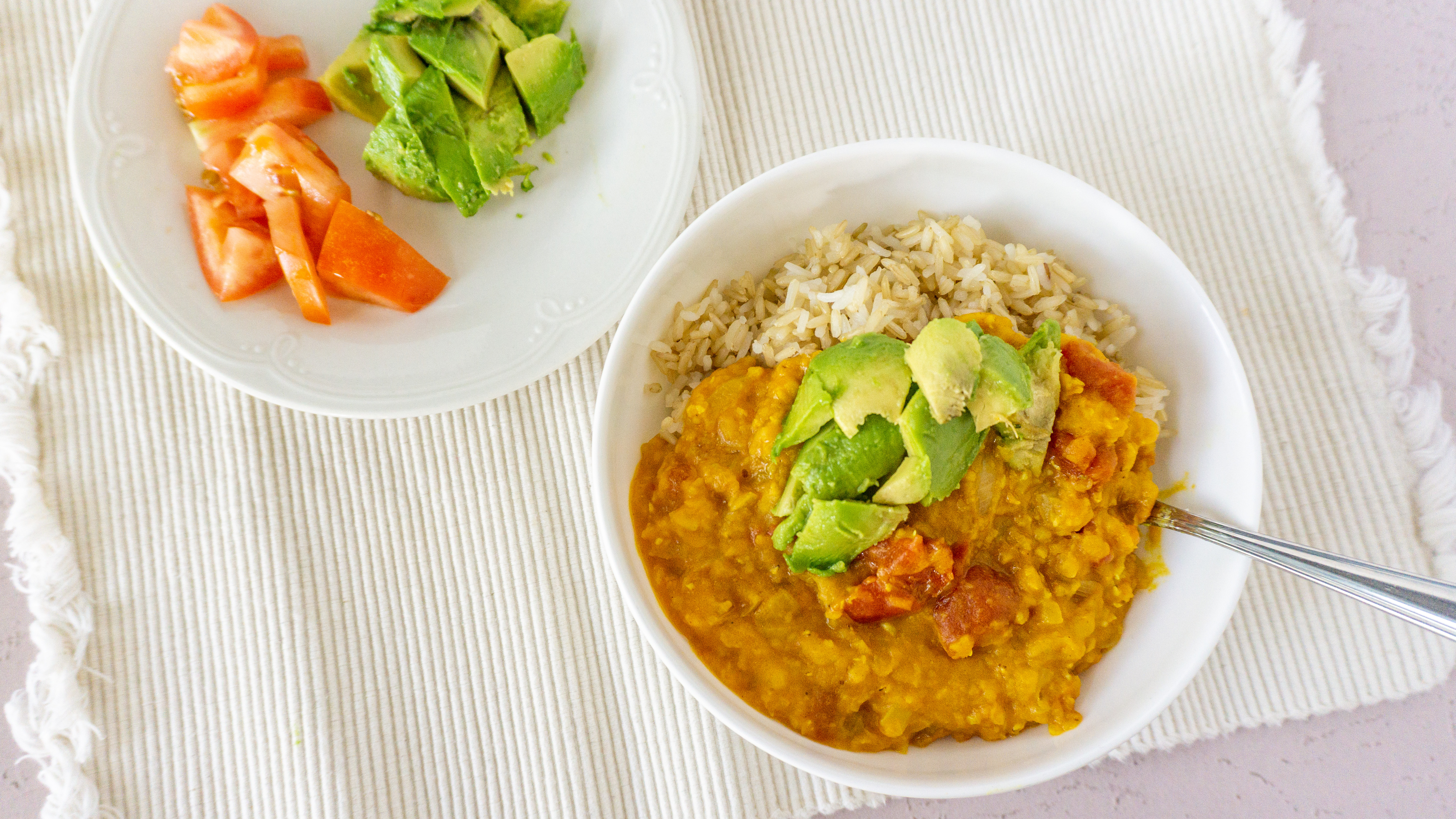 lentil curry on rice in a white bowl with tomatoes and avocado on the side