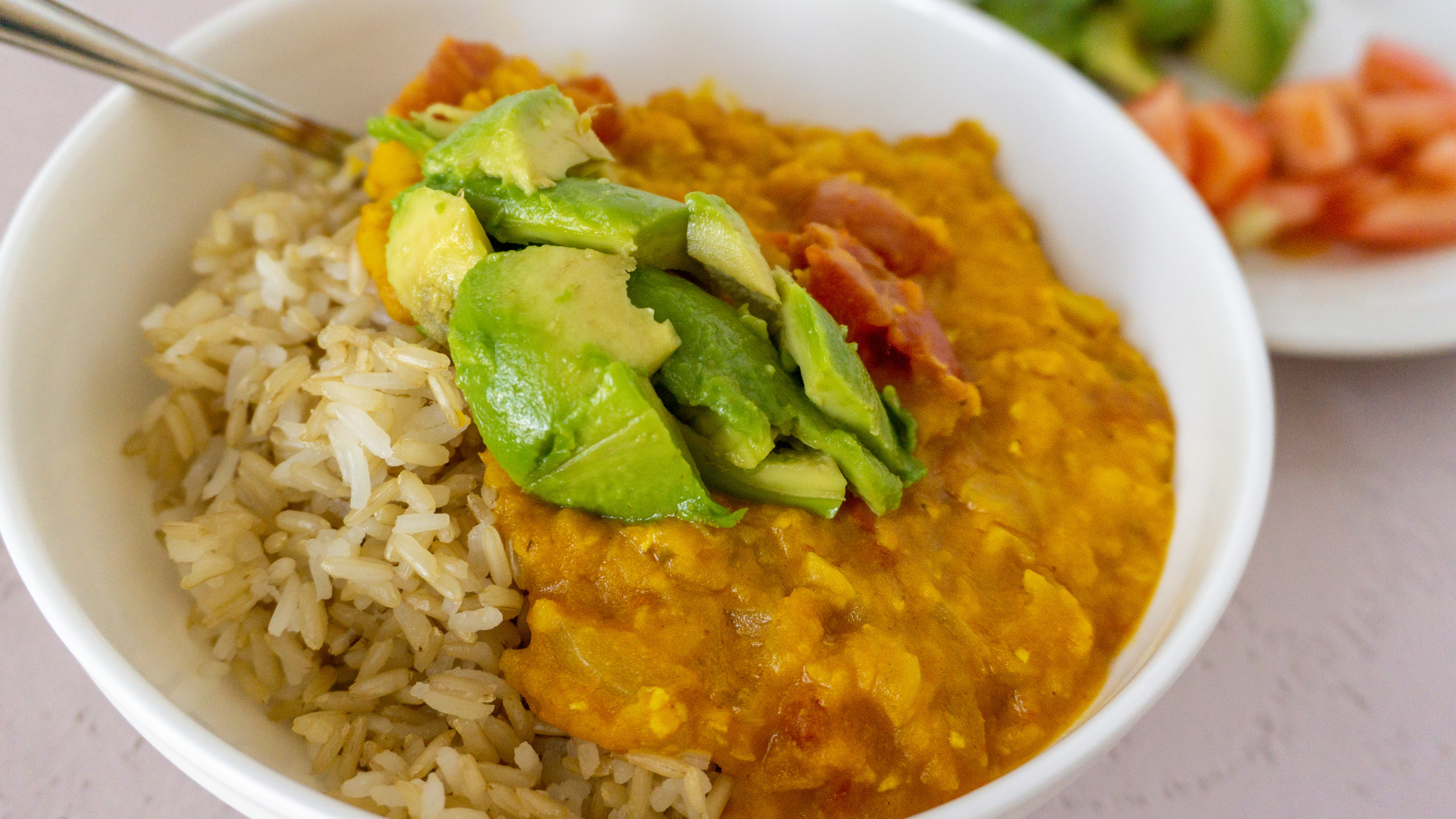 vegan lentil curry on top of rice with avocado on top and tomatoes on the side.